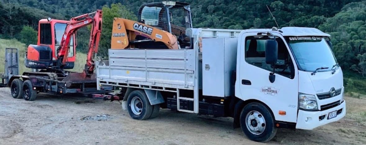 Construction vehicles parked outdoors: red mini excavator and orange loader on flatbed truck, white service vehicle beside them
