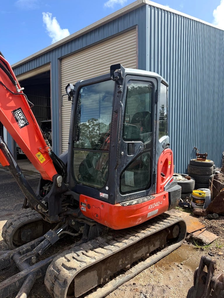 Red and black compact excavator with tracked treads parked outside an industrial metal building on a sunny day