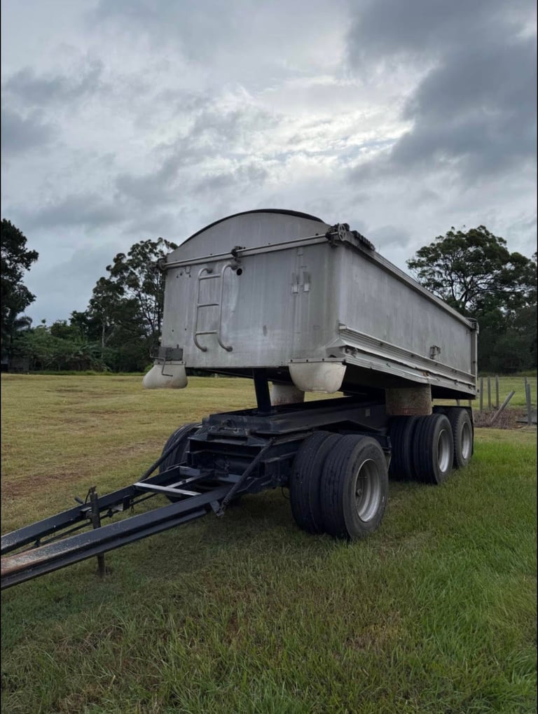 White grain cart trailer with dark chassis parked in a grassy field under cloudy sky, surrounded by trees