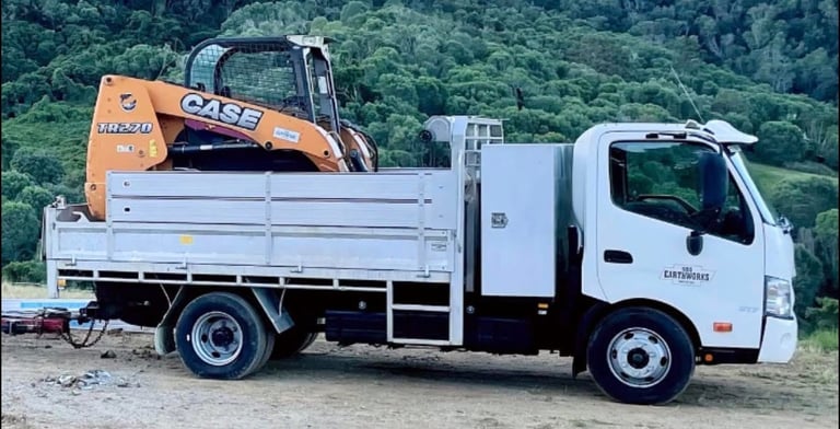 White flatbed truck carrying an orange CASE mini excavator on its bed with forested hills in background